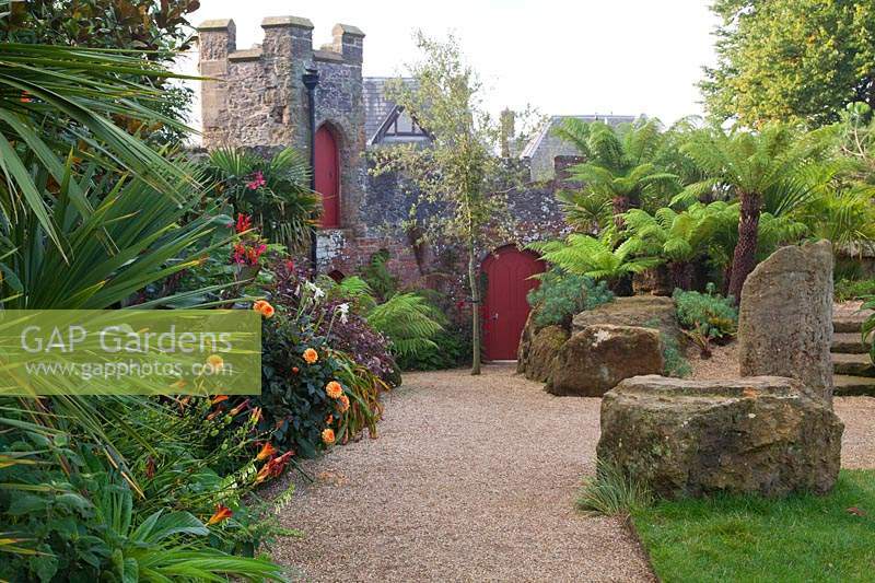 Path alongside large stones, Dicksonia Antarctica - tree fern, Dahlia and 
Canna, to red arched gates in wall 