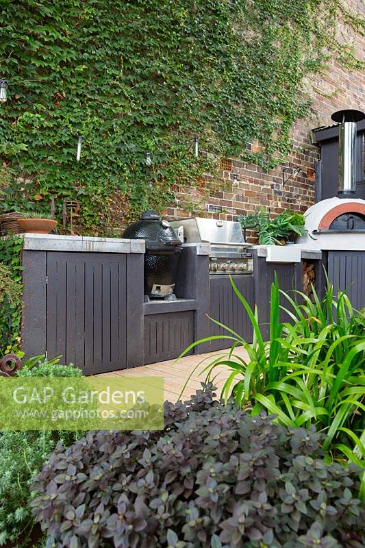 View of outdoor cooking area on terrace patio, with barbecues and pizza oven, surrounded by lush planting. 
