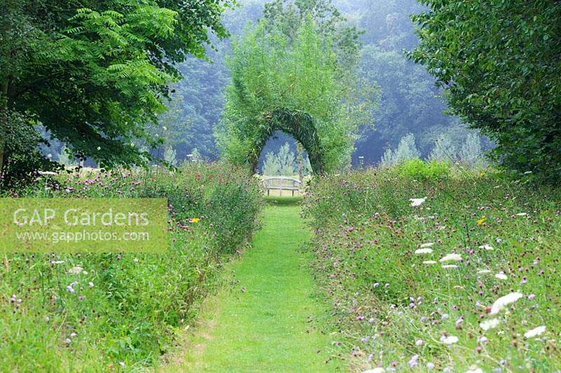 Looking down mown pathway through the wildflower meadow towards the living willow arch and bench in the parkland beyond.