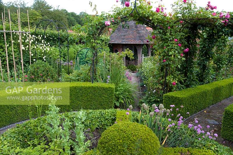 An ornamental kitchen garden with roses climbing over arches, clipped Buxus - box edging and a herb wheel edged with Hedera - ivy