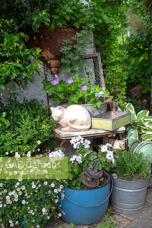 Display of collectables, including a cat ornament, on a chair. Surrounded by planted up vintage buckets.