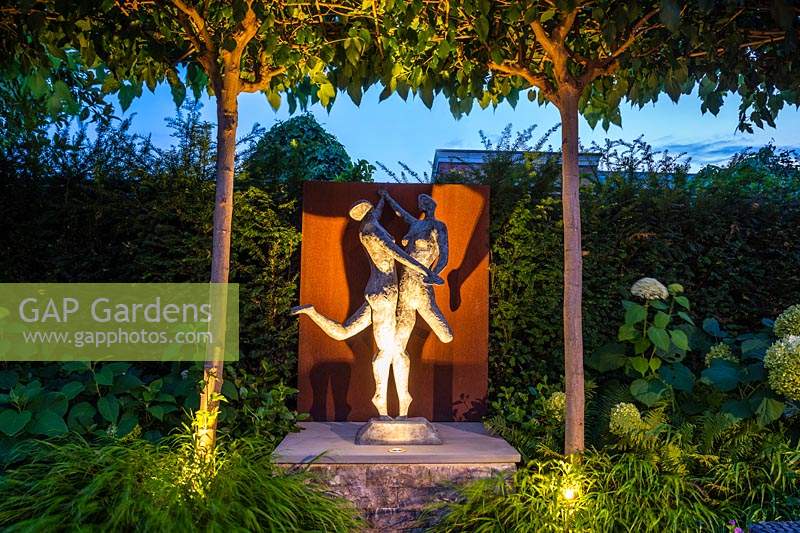 Illuminated statue of dancing couple under canopy of trained Platanus acerifolia - Umbrella Head Tree.
