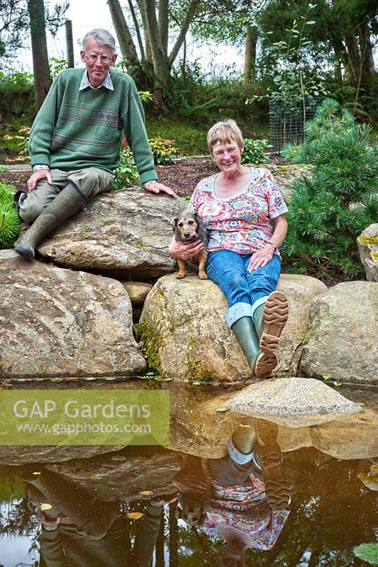 Margaret and Iain Gimblett with pet dog sitting on boulders by the edge of the pond. 
