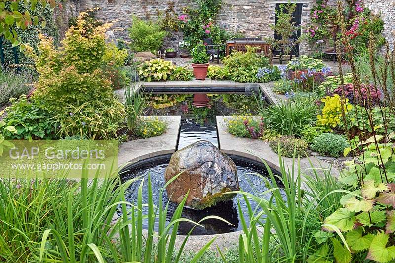 Pond surrounded by flowering borders in walled garden.