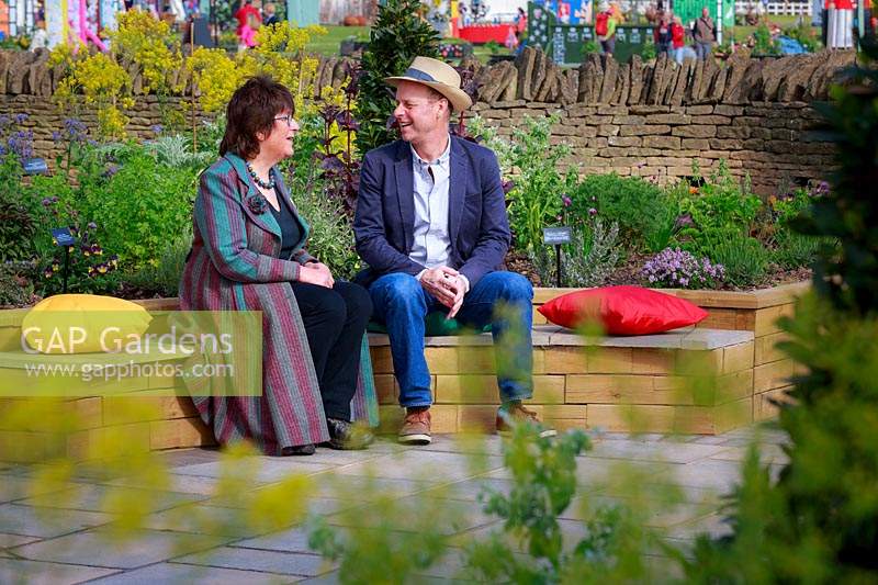 Part of the Health and Wellbeing permanent garden, Jekka McVicar and 
Joe Swift sit in conversation on brightly coloured cushions in the 
central circular sitting area, with herbs in the foreground 
