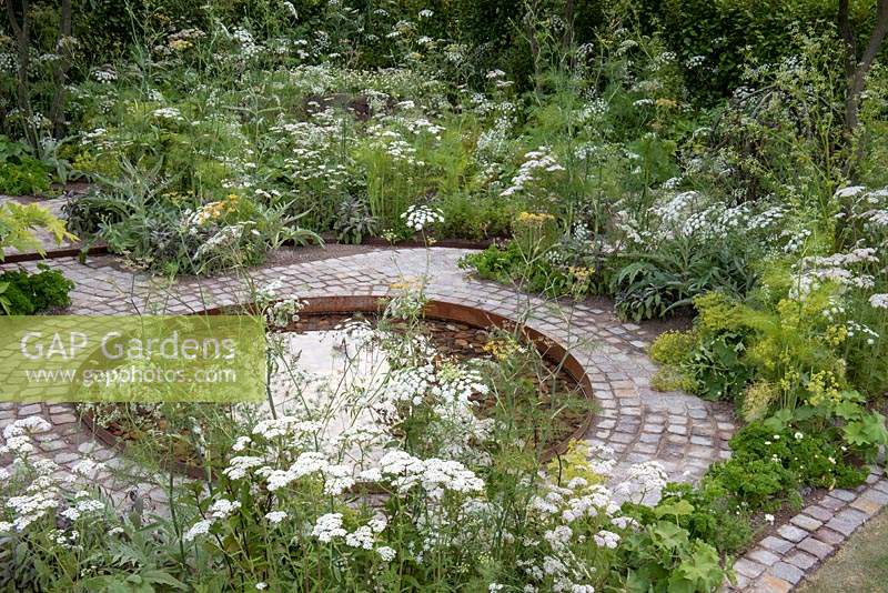 Circular pool edged with a cobbled path and herbs. The Health and Wellbeing Garden, designed by Alexandra Noble, sponsored by CED Ltd, Majestic Trees, Marshal Murray, Hampton Court Palace Flower Show, 2018 
