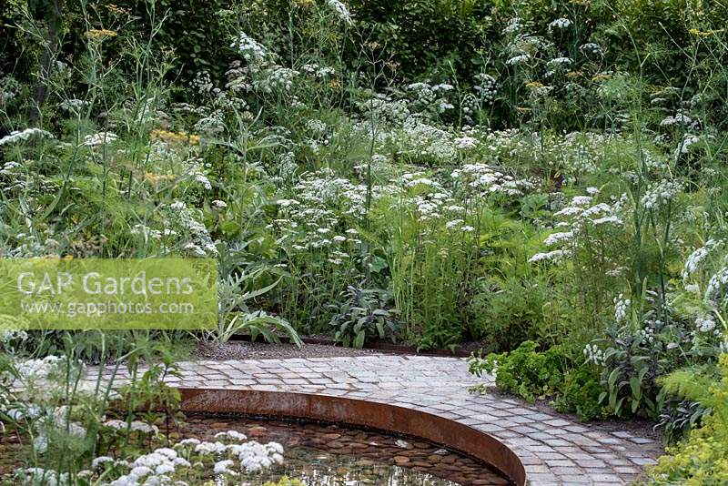 Circular pool edged with a cobbled path and herbs. The Health and Wellbeing Garden, designed by Alexandra Noble, sponsored by CED Ltd, Majestic Trees, Marshal Murray, Hampton Court Palace Flower Show, 2018
