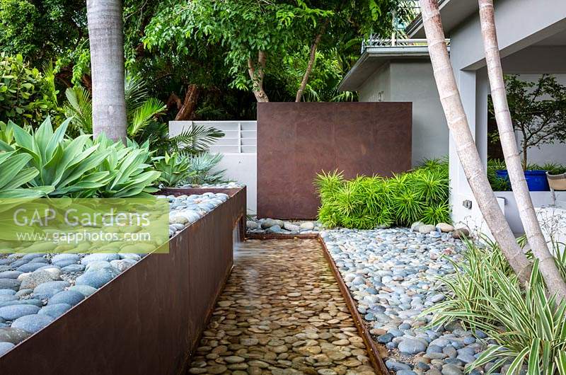 Corten steel water feature with pebbles or cobbles near house, block planting of 
Agave weberi, Osmoxylon lineare - Miagos Bush and Phormium - Variegated Flax
