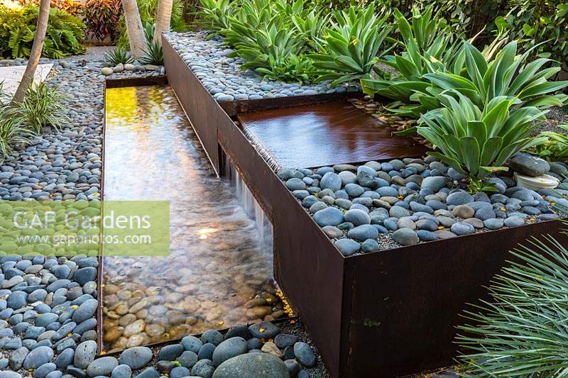 View along a water feature made of corten steel and cobbles that includes a rill, waterfall and planting of 
Agave weberi