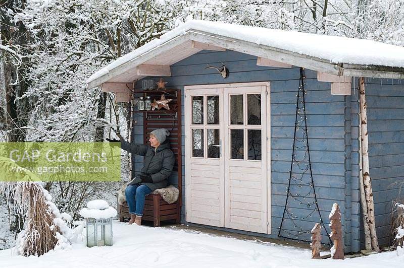 A woman sitting in front of blue summerhouse covered by snow in winter.

