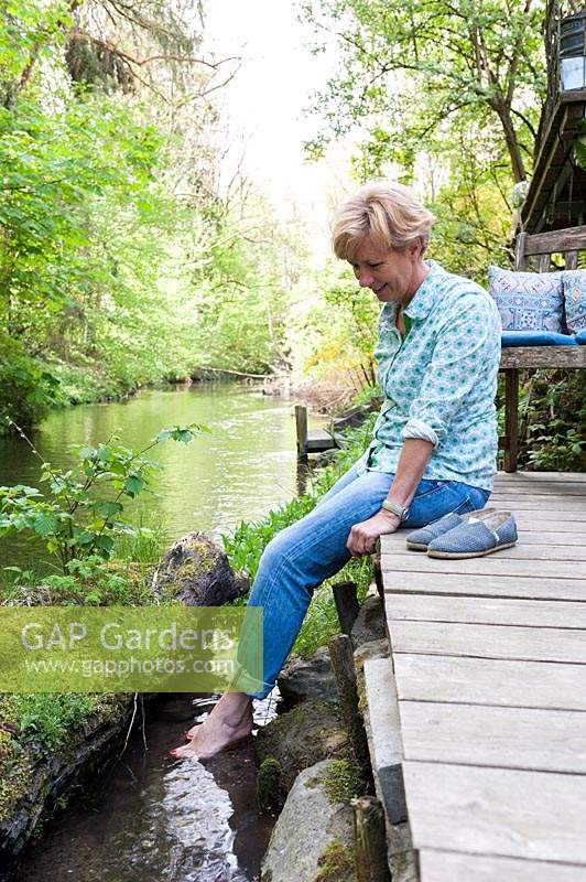 Woman sitting on a deck with her bare feet dangling in the water of a creek
