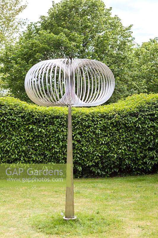 Torodial - sculpture in stainless steel-  on lawn with hedge and trees beyond
