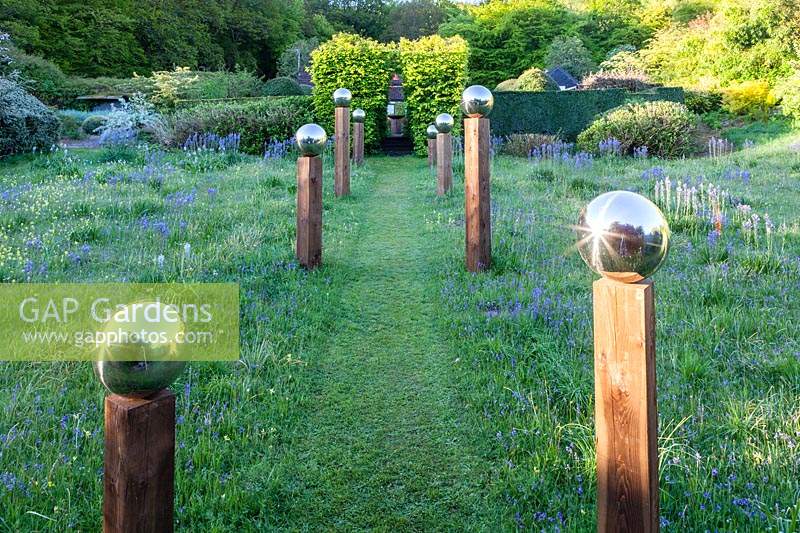 View up grass path in middle of avenue of stainless steel mirror globes
 mounted on wooden posts. Main plant in meadow is Camassia subsp. leichtlinii
