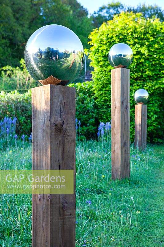 Close up of a stainless steel mirror globe mounted on wooden post, part of an avenue by path in a meadow with
Camassia subsp. leichtlinii
