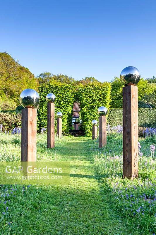 View along grass path with an avenue of stainless steel mirror globes mounted on wooden posts
