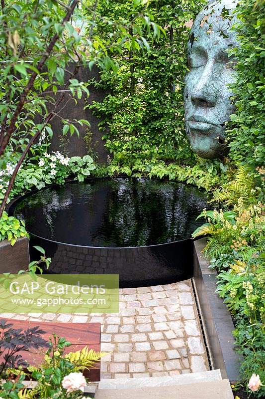 'The Thrive Reflective Mind' garden with raised pond set on paving with sculpture within hedge
