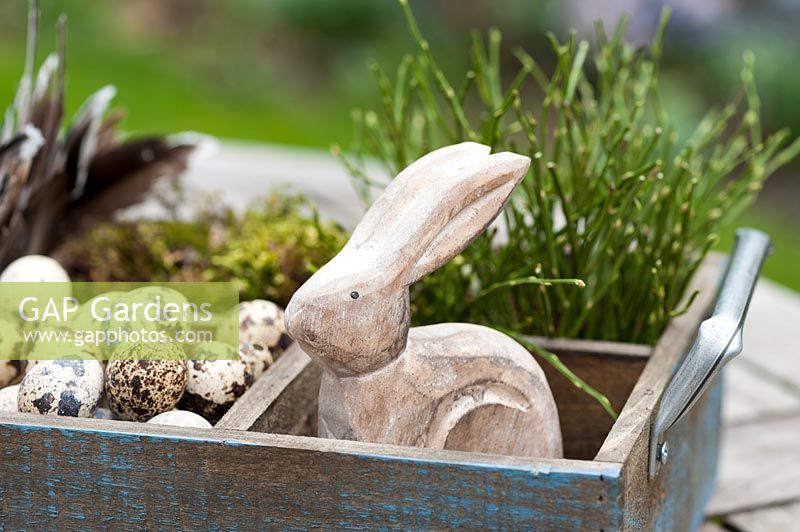 Easter decoration on a table in a spring garden. The wooden box is filled with Viola, quail eggs, a wooden Easter bunny, bilberry branches, moss and feathers.