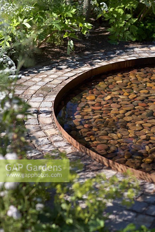 Circular pool with pebbles and corten steel edging. The Health and Wellbeing Garden, designed by Alexandra Noble. Sponsors: CED Ltd, Majestic Trees, Marshall Murray. RHS Hampton Court Flower Show, 2018.
