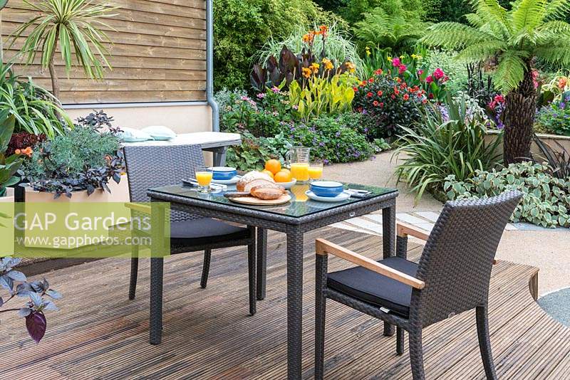 Intimate dining area on a deck overlooking a courtyard edged in beds of Australian tree ferns, cannas, phormiums, dahlias, hardy geraniums and silver Plectranthus.