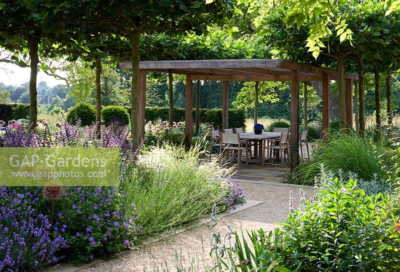 Wooden pergola creates shaded dining area, surrounded by trees, flowering perennials and ornamental grasses. 