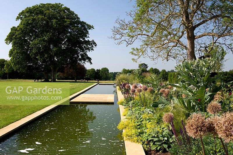 Long, modern water feature by lawn and flowering perennial border.
