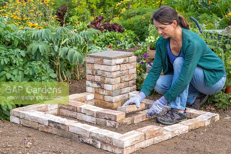 Woman adding bricks to create two L-shaped tiered beds around the corner pedestal. 
