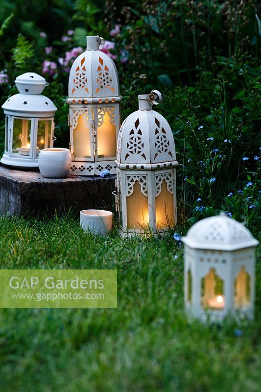 An eclectic mix of white candle lanterns in garden at dusk.