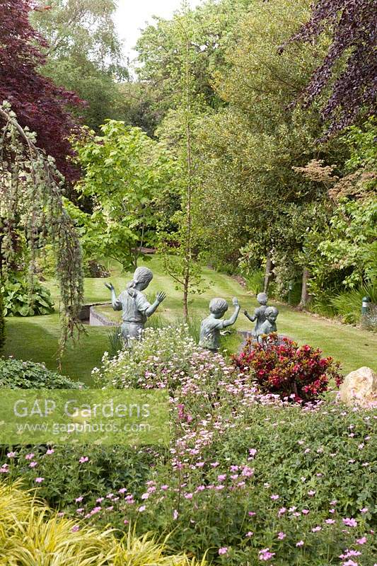 View across herbaceous border to lawn with bronze figures of children in a rill - May