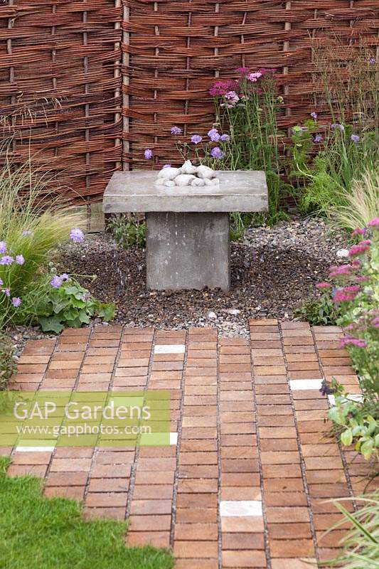 A contemporary stone fountain with woven willow fence and concrete and oak setts paving in the 'Raised By Rivers' Garden at Tatton RHS Flower Show 2018