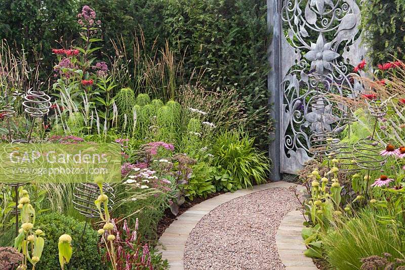 A curved gravel path with stone edges leading through borders to an ornate metal gate with perennial planting including Helianthus salicifolius, Phlomis russeliana and Monarda 'Jacob Cline' in the 'Don't Chop Me Down' Garden at Tatton RHS Flower Show 2018
