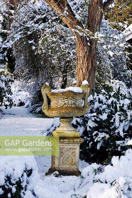 View through a snowy garden with stone urn at Burrow Farm Garden, Dalwood, Devon, UK. 