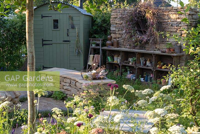 A romantic country garden planted with mostly medicinal plants, a shepherd's hut, a curving stone bench, and an eclectic display of objects. The Naturecraft Garden, designed by Pollyanna Wilkinson, RHS Hampton Court Palace Garden Festival, 2019.