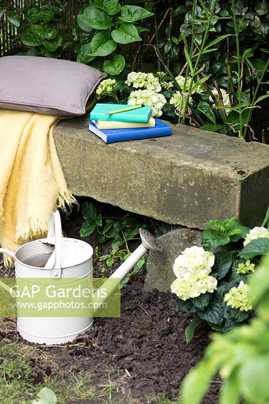 Simple stone bench with cushion, throw, books and watering can