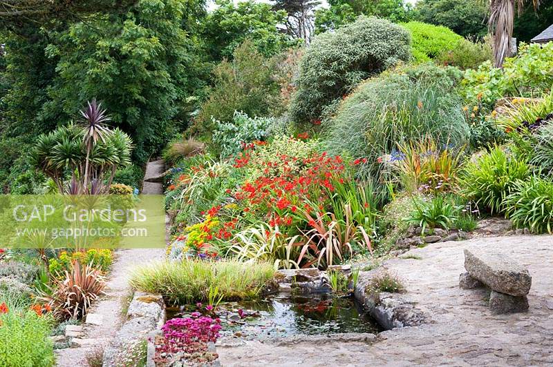 A sunken pond in the corner of the terrace with a path leading between lushly planted sloping borders beyond. Planting includes cordylines, phormiums, Crocosmia 'Lucifer', grasses and euphorbias.