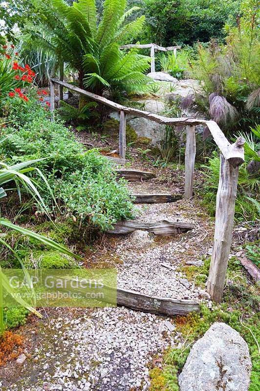 Riven oak handrails snake up the steepiy sloping garden beside shallow steps, past large granite outcrops surrounded by lush ferns including Dicksonia antartica - Tree Ferns. 