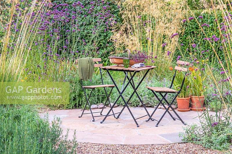 Wooden table and chair bistro set on circular patio, surrounded by ornamental grasses and Verbena bonariensis.
