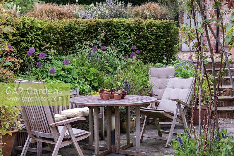 A sheltered seating area, tucked away below a yew hedged and raised beds studded with purple allium and hardy geraniums.