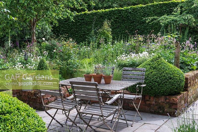 A small paved courtyard with dining table and chairs, flanked by domes of box, overlooks a box parterre planted with Bistort, hardy Geraniums, Centaurea, Foxgloves, ragged robin, Astrantias, Aquilegias and Roses.
