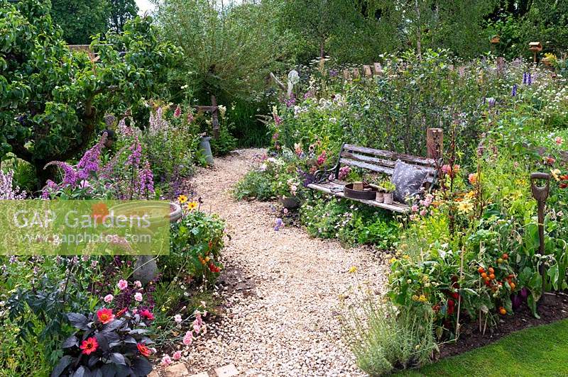 Gardening tools and cushion on rustic garden bench nestled amongst perennial planting along winding gravel path. RHS Hampton Court Festival 2019.
