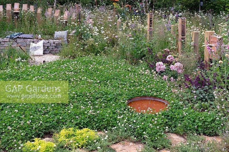 Clover lawn with small pond and curved dry stone bench backed with timber palisade, fencing posts with insect hotels in semi wild garden. RHS Hampton Court Festival 2019.