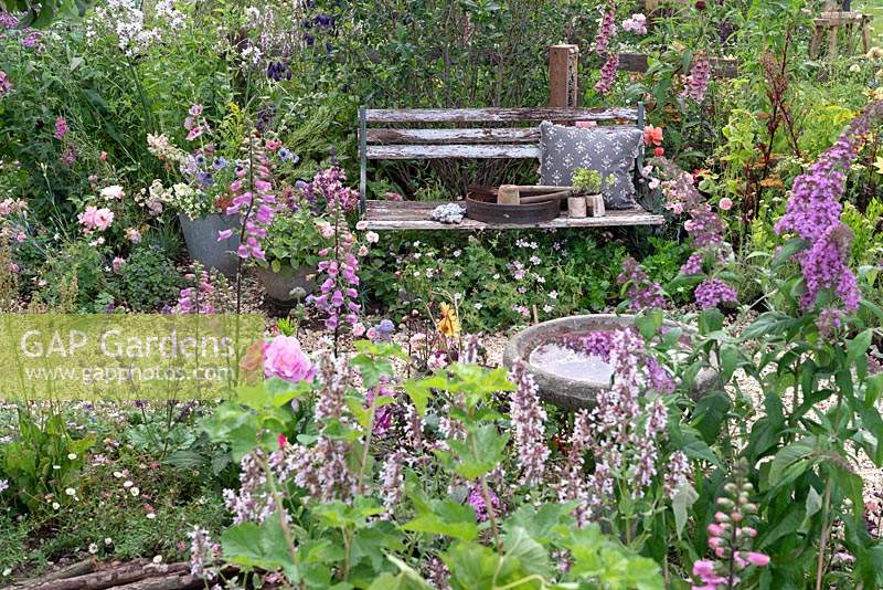 Gardening tools and cushion on rustic garden bench nestled amongst perennial planting along winding gravel path. RHS Hampton Court Festival 2019. 