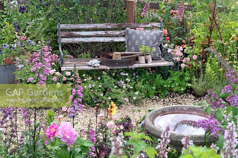 Gardening tools and cushion on Rustic garden bench nestled amongst perennial planting along winding gravel path. RHS Hampton Court Festival 2019.