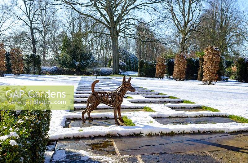 Sculpture of terrier by Harriet Mead, made from reclaimed metal machinery. Carpinus betulus - Hornbeam pillars, Taxus baccatta - yew hedge with stone paving and Fagus sylvatica f. purpurea - large copper beech.  Buxus - box hedging. Snow in January.