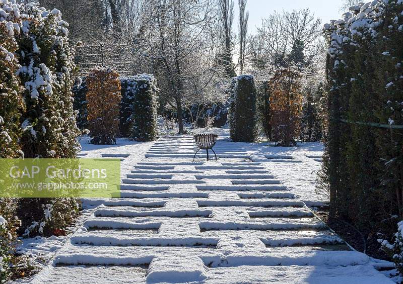 Carpinus betulus - Hornbeam pillars, Taxus baccata - yew hedge with stone paving and small metal fire pit. Snow in January.