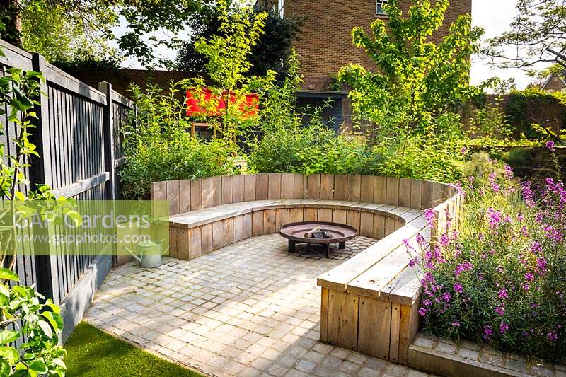 Seating area with fire pit and round wooden bench surrounded by Erysimum 'Bowles's Mauve', Prunus avium 'Stella' and Cornus alba 'Sibirica' .