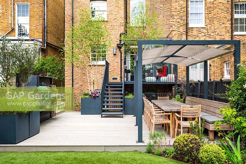 Modern outdoor kitchen with BBQ and dining area and big cubic containers with Olea europaea Olive trees, Salvia officinalis and Betula pendula multi-stem underplanted with Heuchera x 'Plum Pudding'