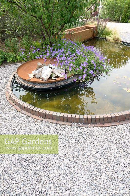 Round pond edged with bricks set in gravel. Within the pond a fire pit with Geranium 'Rozanne'