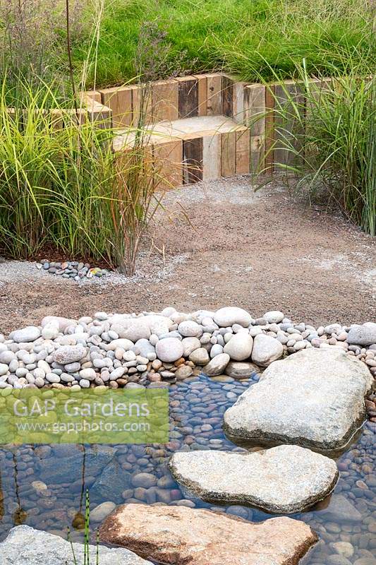 A naturalistic style garden using natural materials - a shallow pond with pebbles and stepping stones, and a wooden seat set into a sloping garden with grasses. RHS Hampton Court Palace Garden Festival 2019.