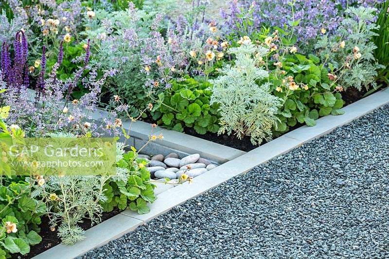 Use of stone in the garden - a grey gravel path is neatly edged with a contemporary style white stonework and pebbles, with blue and silver plants. RHS Hampton Court Palace Garden Festival 2019. Sponsor: Viking Cruises