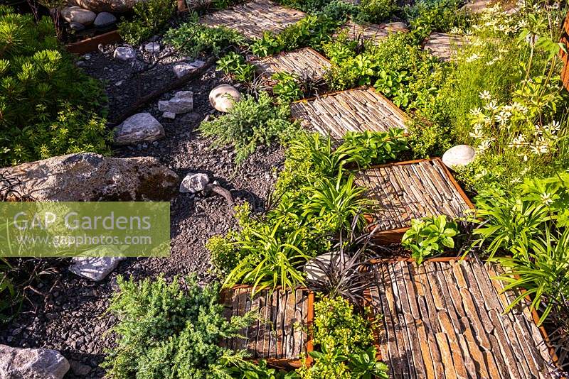 Rusted corten steel encases square blocks of rough cut natural stone, forming a path through the a green foliage in the Through Your Eyes garden at the RHS Hampton Court Palace Garden Festival 2019. Sponsors: Kebony, CED Stone, R and G Metal Products, William's Art and Design and Practicality Brown